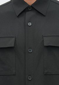 Mustast kandimiskinnisest särgist, millel on kaks esesahtlit. Omadusteks on struktureeritud krae, sile pind ja mustad nupud. Lihtne ja funktsionaalne disain.