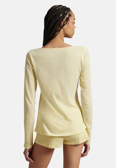 Femme aux cheveux tressés portant un haut à manches longues jaune pâle et un short assorti en tricot côtelé, vue de dos sur un fond blanc.