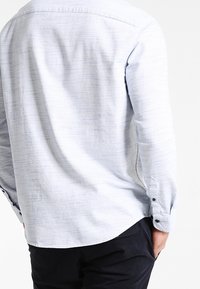 Chemise bleu clair à manches longues avec une texture subtile, vue de dos. Elle présente un col boutonné et des boutons foncés sur les poignets, avec un ourlet droit.