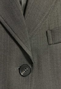 Gros plan d'un blazer en tissu gris texturé avec un bouton noir à quatre trous et une petite poche poitrine sur le côté droit.