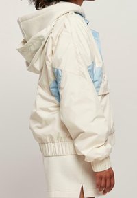 Chaqueta ligera de color crema con acentos azules, cuello alto y puños elásticos. Presenta una capucha en contraste y un diseño de corte relajado.