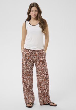 Žena s dugom smeđom kosom, odjevena u bijelu majicu bez rukava i široke hlače s cvjetnim uzorkom, s rukama u džepovima, stoji na jednostavnoj pozadini.