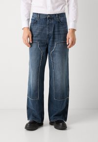 Široke denim hlače z nasprotnimi zaklopkami, svetlo in temno modre odtenke, raztrgane robove in sproščen kroj. Črna obuvala se dopolnjujejo.