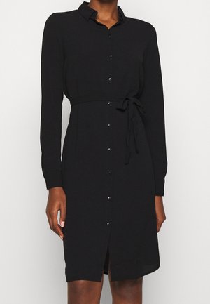 Czarna sukienka na guziki z długim rękawem, kołnierzykiem i wiązaniem w talii, noszona przez osobę o ciemnej karnacji, stojącą na jednolitym jasnym tle.