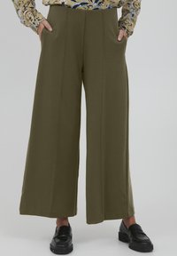 Pantalons larges couleur khaki en tissu doux, avec des poches avant, une finition lisse et une coupe ajustée. Portés avec des chaussures noires.
