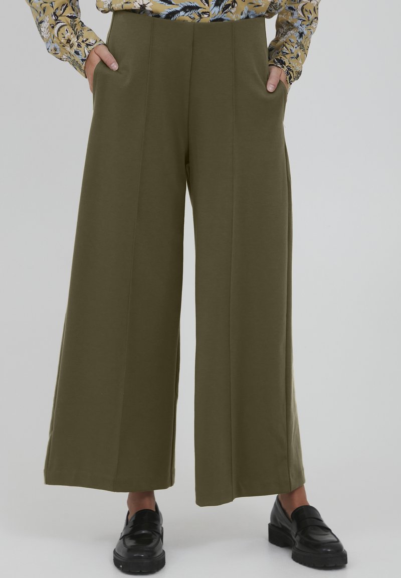 Pantalons larges couleur khaki en tissu doux, avec des poches avant, une finition lisse et une coupe ajustée. Portés avec des chaussures noires.