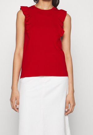 Czerwona bluzka bez rękawów z falbaniastymi akcentami na ramionach; miękka tkanina; okrągły dekolt; noszona z białą spódnicą, prezentując luźny krój.
