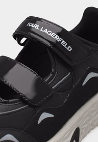 Μαύρο αθλητικό παπούτσι Karl Lagerfeld με γυαλιστερές ρυθμιζόμενες λουρίδες και λευκή λεπτομέρεια μοτίβου στη σόλα που είναι ορατή.