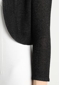 Bolero negro de punto con una textura suave, que presenta un borde redondeado y mangas de tres cuartos, diseñado para complementar diversas prendas.