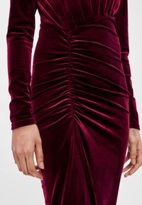 Βελούδινο μπορντό φόρεμα με μακριά μανίκια, που διαθέτει ρυτιδωτές λεπτομέρειες κατά μήκος της πλευράς, δημιουργώντας μια υφασμάτινη και εφαρμοστή σιλουέτα.