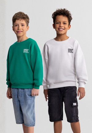 Dwóch uśmiechniętych chłopców stoi obok siebie; jeden ma na sobie zieloną bluzę i dżinsowe szorty, drugi białą bluzę i czarne szorty typu cargo.