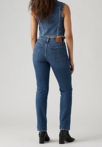 Žena stojící zády, oblečená v modrých džínech s vysokým pasem, bezrukém zkráceném denimovém kabátě a černých vysokých botách na podpatku.