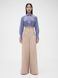 Camicia blu chiaro a righe, cropped, con colletto; pantaloni beige a gamba larga con bottoni decorativi e una texture liscia.