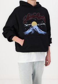 Svart hoodie med framficka, med en färgglad grafik av berg och sol. Mjuk textur, oversized passform, ribbade ärmslut och nederkant.