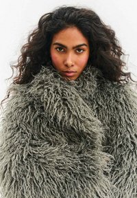 Kobieta o długich kręconych ciemnych włosach, ubrana w gruby, teksturowany szary futrzany płaszcz, patrząca prosto w kamerę z neutralnym wyrazem twarzy.