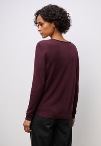 Maglione leggero color bordeaux con maniche lunghe e polsini a coste. Presenta un ampio scollo e una finitura liscia e testurizzata.