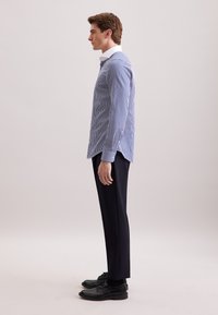 Giovane uomo in camicia a righe blu e bianche, pantaloni neri e scarpe eleganti nere in profilo contro uno sfondo semplice.