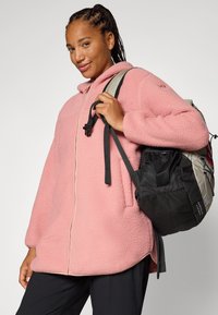 Eine lächelnde Person mit geflochtenem Haar trägt eine rosa Fleecejacke und hat einen schwarzen Rucksack mit einem grauen Gurt auf der Schulter.
