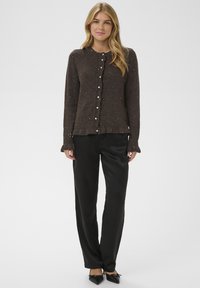 Cardigan marron texturé avec encolure ronde, boutons en perle sur le devant et ourlet à volants, associé à un pantalon en satin noir et des ballerines pointues.