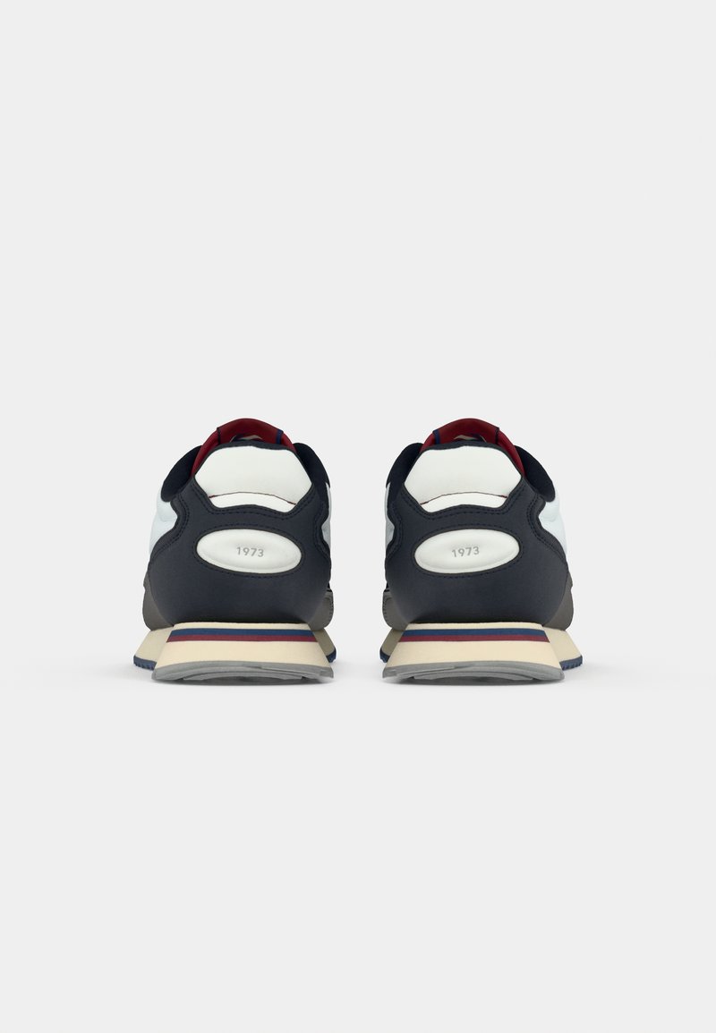 Zapatillas azul marino con una parte superior de malla blanca y gris, acentos rojos, media suela de espuma y tira de sujeción trasera. Presentan un diseño retro con rayas horizontales.