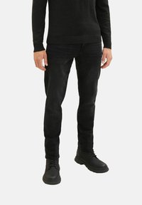 Mand iført sort sweater, sorte slim-fit jeans og sorte snørestøvler står mod en ensfarvet baggrund.