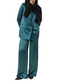 Satynowy garnitur w kolorze morskim z dwurzędową marynarką i szerokimi spodniami, zestawiony z czarnym prążkowanym szalikiem. Gładka tekstura i błyszczące wykończenie.