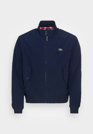 Marineblauwe zip-up jack over een marineblauw polo, beige broeken en blauwe sneakers. Een marineblauwe tas met een groen logo wordt in één hand vastgehouden.