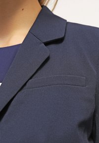 Náměsíčně modrý blazer s hladkou texturou, s klopovým límcem a kapsou na hrudi na levé straně. Jednoduchý a přiléhavý design.