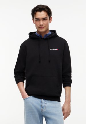 Jonge man die een zwart Tommy Jeans hoodie draagt over een blauw geruit overhemd en lichtblauwe spijkerbroek, staand met één hand in zijn zak.