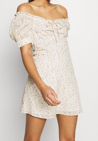 Kobieta w krótkiej, kwiatowej sukience bez ramiączek z bufiastymi rękawami i detalem w postaci węzła z przodu na jednolitym tle.