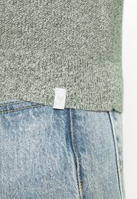 Suéter tejido gris-verde con patrón texturizado y dobladillo acanalado, combinado con jeans de mezclilla azul claro con detalle visible de costuras.