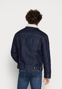 Mörkblå denimjacka med vit shearlingkrage, synlig stickning och knappdetaljer. Stylad med blå jeans, sedd från baksidan.