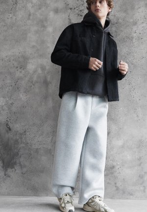 Young person stands against concrete wall wearing black jacket, dark hoodie, light gray wide pants, and white sneakers.