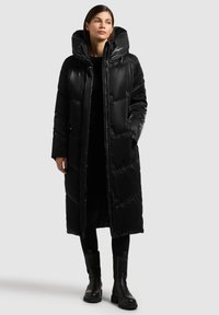 Zwarte donsjas met een lange silhouet en capuchon. Heeft een glanzende textuur, ritssluiting en zijzakken. Draagt dikke zwarte laarzen.