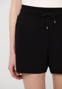 Pantalones cortos de algodón negros con un diseño liso, cintura con cordón y acentos metálicos. Textura suave y un ajuste relajado, llegando hasta la mitad del muslo.