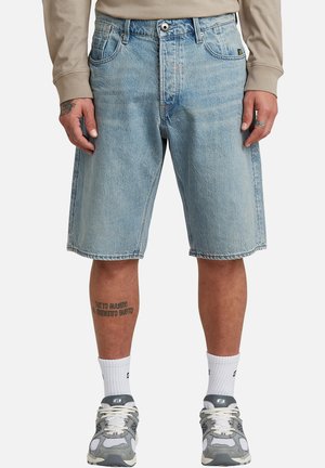 Lichtblauwe denim knielange shorts gedragen met een beige top met lange mouwen, witte sokken en grijze sportschoenen bij een staand persoon.