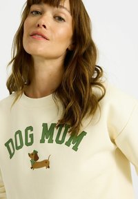 Sudadera de color crema con texto verde que dice "DOG MUM" y un dachshund bordado con pajarita. Tela suave y texturizada.
