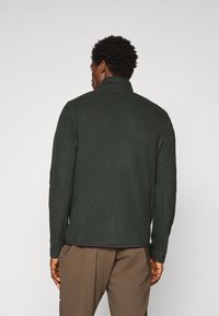 Homme portant une veste en polaire texturée vert foncé et un pantalon marron, vu de dos sur fond clair.