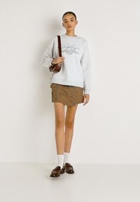 Pelēks sporta krekls ar uzdrukātu tekstu, brūna svītraina mini svārciņa, balti zeķīši un tumši brūnas kurpes bez šņoru. Modele pozē ar brūnu plecu somu.