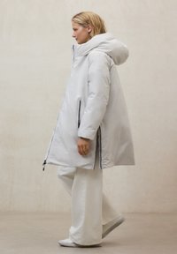 Abrigo largo acolchado de color gris claro con capucha. Cuenta con cremalleras negras y bolsillos laterales. Textura lisa y ligeramente brillante. Diseño minimalista.