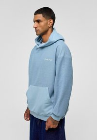 Lichtblauwe hoodie met een textuurstof, een grote voorzak en een relaxte pasvorm. Subtiel geborduurd logo op de borst.