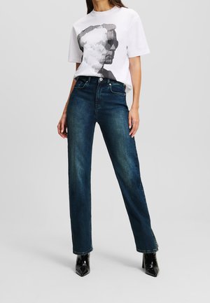 Vrouw draagt een wit grafisch t-shirt met een zwart-wit gezichtprint, donkerblauwe hooggetailleerde jeans en zwarte puntige laarzen.