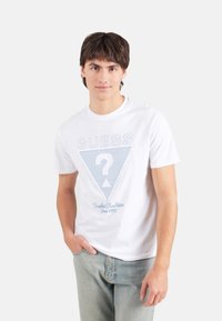 Camiseta de algodón blanca con un logo de triángulo azul que tiene un signo de interrogación y el texto "Tradiciones Atemporales Desde 1981" abajo, de mangas cortas.