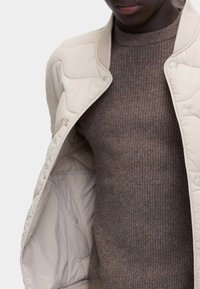 Ljusbeige kuddad bomberjacka med knappstängning, kombinerad med en texturerad brun ribbad tröja. Exponerad insida visar en slät foder.