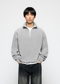 Mann iført en lys grå half-zip strikkegenser over en hvit skjorte og falmede svarte jeans med hendene i lommene mot hvit bakgrunn.