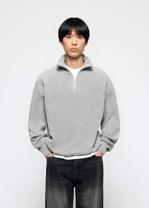 Mann in einem hellgrauen Strickpullover mit halbem Reißverschluss über einem weißen Hemd und verblichenen schwarzen Jeans, mit den Händen in den Taschen, vor einem weißen Hintergrund.