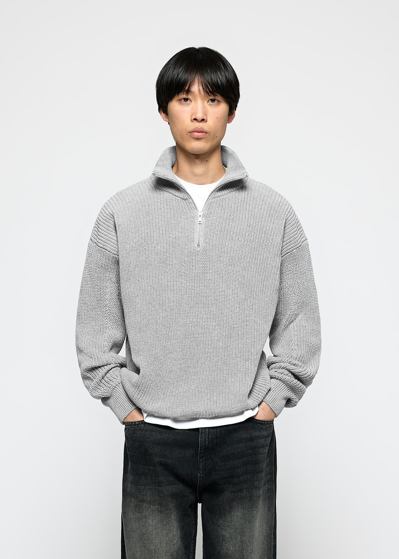 Mann iført en lys grå half-zip strikkegenser over en hvit skjorte og falmede svarte jeans med hendene i lommene mot hvit bakgrunn.