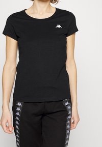 Černé tričko s krátkým rukávem vyrobené z bavlny, s kulatým výstřihem a malým bílým logem na levé straně hrudi.