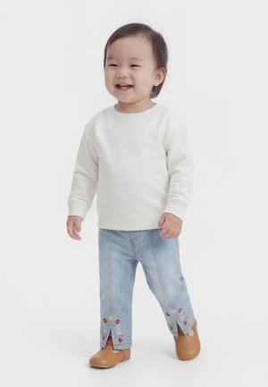 Malo dete se smehlja, nosi belo majico z dolgimi rokavi, svetlo modre kavbojke z rožnim vezenjem in rjave čevlje, stoji na belem ozadju.