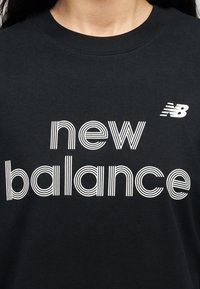 Μαύρο μπλουζάκι με λαιμόκοψη, με λευκή ρίγα και το κείμενο "new balance" καθώς και ένα μικρό λογότυπο "NB" στο πάνω δεξί μέρος του στήθους, που φοράει ένα άτομο.